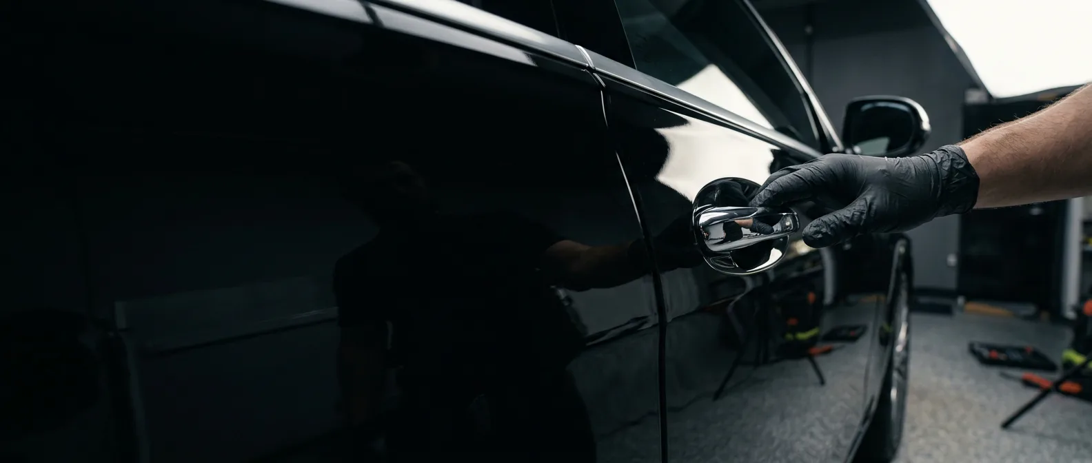 Gloved hand on the chrome door handle of a glossy black luxury car under soft studio lighting at Brilliant Shine